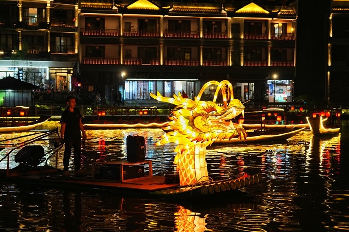 2025年10月5日晚，中秋节将至，湖南凤凰古城沱江上，百米灯光金龙游船拼接出圆月图案。金龙游船首尾相接，与两岸吊脚楼灯火相映，吸引游客沉浸式感受古城中秋氛围。图为船员驾驶金龙游船在河中行驶。