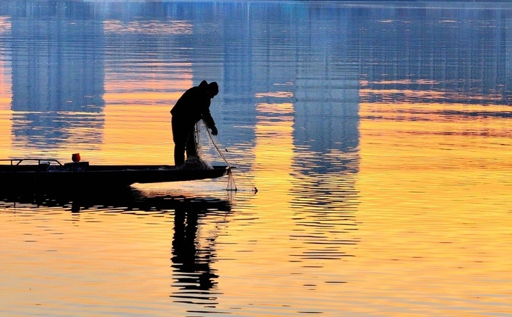 2025年10月24日，黑龙江省哈尔滨市，初冬的清晨，松花江水平如镜，倒映着天边微光。一艘艘小渔船轻划江面，渔民们娴熟地起网、下网，银丝渔网在晨光中闪烁，网上跃动的鱼影与波光交织。金黄的晨光铺满江面，将渔船、渔网和渔民的剪影染成温暖的金色，宛如一曲无声的渔光曲。江面倒影摇曳，渔船与天际线融为一体，构成一幅静谧而生动的初冬捕鱼图。IC photo