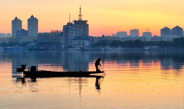 2025年10月24日，黑龙江省哈尔滨市，初冬的清晨，松花江水平如镜，倒映着天边微光。一艘艘小渔船轻划江面，渔民们娴熟地起网、下网，银丝渔网在晨光中闪烁，网上跃动的鱼影与波光交织。金黄的晨光铺满江面，将渔船、渔网和渔民的剪影染成温暖的金色，宛如一曲无声的渔光曲。江面倒影摇曳，渔船与天际线融为一体，构成一幅静谧而生动的初冬捕鱼图。
