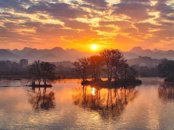 2025年12月15日，广西桂林，太阳从远处的山顶渐渐升起，温暖的阳光照耀着远处的山峦和村庄，映红了湘江，江面上的小舟在收拾渔获，码头广场还有晨练的瑜伽…冬日暖阳晨景美不胜收。