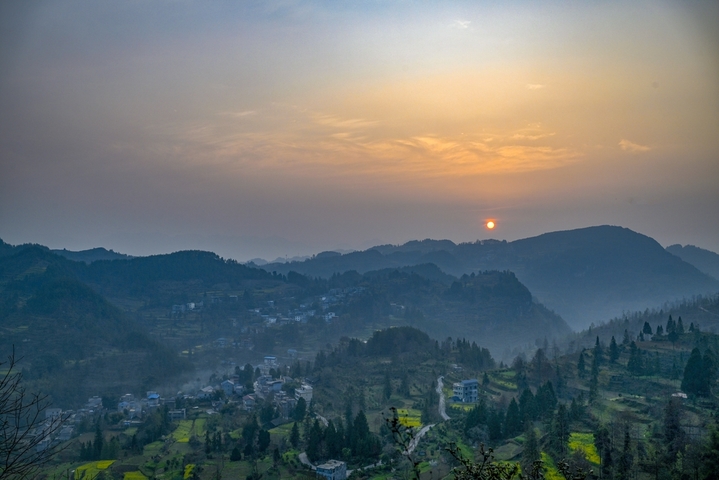 2026年3月13日，重庆，酉阳土家族苗族自治县丁市镇沙溪村，红日跃出群山，薄雾轻笼村落。蜿蜒山道串起层层梯田，袅袅炊烟随霞光升腾，与田间新绿、林间繁花相映。晨光漫过青瓦屋舍，唤醒沉睡的山乡，勾勒出一幅充满烟火气的渝东南春日晨景，尽显乡村田园的诗意与生机。